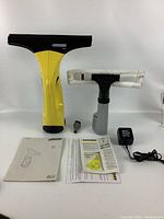 Front view of Karcher WV 50 window vacuum and accessories including spray bottle with microfiber pad, charger, brush nozzle, instruction manuals, and cleaning items.