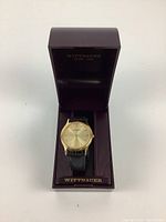 Front view of the Wittnauer watch in original open box showing gold-tone case, black leather strap and burgundy interior box.
