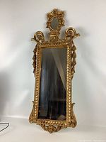 Front view of narrow rectangular mirror with ornate gilted frame and decorative top with oval mirrored inset.