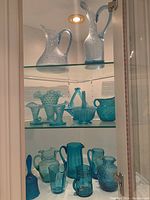 Full view of three glass shelves displaying various blue glassware items including hobnail pitchers, vases, bell, mug, and jugs.