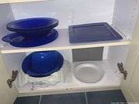 Cabinet shelf showing blue glass rectangular platter stacked with eight large blue glass pasta bowls and a clear round glass plate below