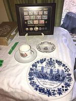 Framed Canada Centennial stamps and coins in wooden frame on stand, with cup and saucers on cloth beneath.