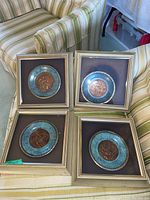 Four framed decorative plates showing copper-tone center with cherubic motifs and blue textured border, arranged on striped fabric surface, one plate has slipped inside frame.