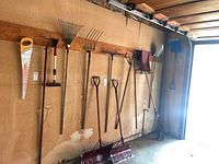 Full view of all garden tools hanging on garage wall including saw, rakes, pitchforks, shovels, snow shovels and squeegee.
