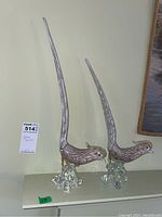Overview photo of two tall Murano glass pheasant bird sculptures side by side on a shelf against a wall, showing full height and posture with long tails