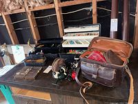 Overview of two tackle boxes (white and black), six reels, brown tackle bag on table