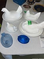 Six chicken-shaped containers on a table; four are white milk glass roosters/hens with lids; two are blue and cobalt blue glass hens with lids.