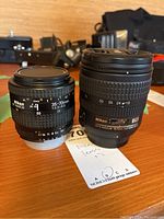 Two Nikon camera lenses side by side on a wooden surface, showing both lens markings and labels.