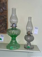 Two antique glass oil lamps side by side, one larger green lamp and one smaller clear etched lamp, standing on a white surface.