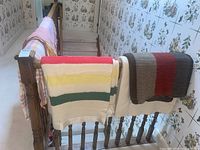 Multiple blankets and quilts hanging over wooden bannister staircase, showing a variety of wool blankets and throws including the Hudsons Bay-style wool blanket with red and brown wide stripes and a multi-colored striped cream blanket