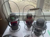 Three vintage railway lanterns on window sill showing red and clear glass globes, metal protective cages, and wire handles.