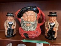 Two small Winston Churchill ceramic pitchers flanking a larger Falstaff ceramic pitcher on a wooden shelf, showing scale and condition