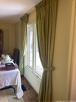 Two green silk drape sets hanging on white wall and window with green tiebacks securing one panel.