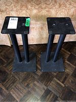 Two black metal speaker stands side by side in front of a couch, viewed from front and top angle.