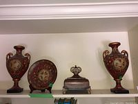 Front view of the two red vases, decorative plate in the middle, and metal trinket box in center on shelf, showing overall set