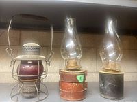 Wide shot showing three vintage train lanterns on a shelf, two with glass chimneys and one with a red glass globe covered by a metal cage