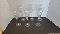 Six clear crystal champagne flutes displayed on a black surface, showing etched patterns and slender stems.