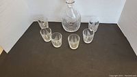 Photo of etched crystal wine decanter with six matching glasses arranged in a semicircle on a black surface against a white backdrop.