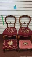 Front view of two matching antique French Provincial style accent chairs with carved wooden frames, curved backs, and red needlepoint upholstered seats with floral designs, placed side by side with two small needlepoint foot stools featuring different floral upholstery patterns in front.