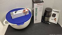 Showing all items together: large white Tupperware bowl with blue lid, boxed black can opener on top, boxed mint green KitchenAid hand blender, and Cuisinart grinder with manual.