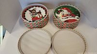 Two round decorative storage boxes with Christmas designs and plate holders inside.