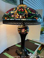 Upper portion of lamp lit showing stained glass shade with floral and leaf motifs and metal lattice trim