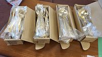 Four boxes containing gold plated forks, spoons, and serving spoons individually wrapped in plastic