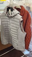 Display of two Arctic Expedition sleeveless vests on hangers: one gray and partially visible red jacket behind them.
