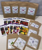 Photo showing 4 brown kraft paper seed pouches labeled 'Sensation Mix', assorted colorful seed envelopes with images of various flowers including Hollyhock, Maltese Cross, Pansy, Poppy, Zinnia, Livingstone Daisy, Marigold, and Italian Large Leaf Basil seed pouch.