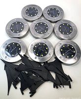 Eight stainless steel circular solar LED lights with black solar panels and embedded LED lights, accompanied by seven black triangular plastic stakes for ground mounting.