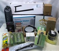 Photo showing multiple boxed houseware items including a water filter, paper towel holder, green glass cups, LED light bulb, garbage bags, candle warmer, and other small boxed and bagged items.