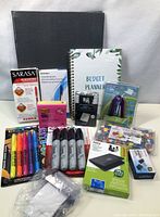 Photo showing array of colorful markers, black Sharpie markers, budget planner, small stapler, and some boxed items of office supplies on white surface.