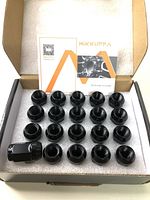 Open box with 20 black alloy lug nuts arranged and a booklet inside the box.