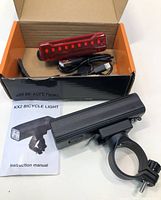 Photo of black rechargeable front bike light with mounting bracket, red rear light, USB charging cable, instruction manual, all inside an open box.