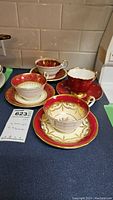 Four Aynsley China teacups with matching saucers showing red, gold and floral designs, placed on a counter.
