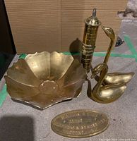 Photo showing all brass items in the lot on a concrete floor including scalloped bowl, two swan figurines, cylindrical brass piece, and oval plaque.