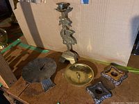 Five metal candleholders arranged on a wooden surface with a cardboard background. Includes tall ornate brass candleholder with floral motif, one round brass dish candleholder, one round silver-toned dish, and two square silver-toned candleholders.