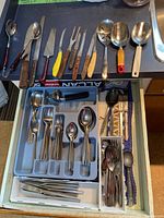 Top edge view of drawer showing wooden-handled spreaders, forks, knives and plastic-handled serving spoons placed on counter above drawer
