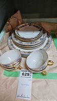 Porcelain dinnerware set including stacked plates and two tea cups with gold handles, placed on brown packing paper.