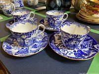 Four Royal Crown Derby porcelain tea cups displayed on matching saucers, featuring blue and white Asian-inspired scenic patterns and gold trim.
