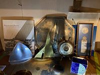 Overall view of the lot showing brass sailing boats, barometer clock, octagonal metal wall clock, framed medal, travel alarm clock, book, and metal decor tray.