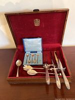 Red velvet-lined wooden box open showing 5 sterling sugar spoons, 2 EPNS salad servers, carving knives and fork.