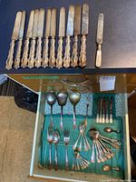 Photo showing 11 silver handled serving knives with ornate floral handles, and a smaller knife with mother of pearl handle on top edge of drawer. Below in drawer are serving spoons, forks and other cutlery including a Sterling silver Birks baby spoon and fruit cutters with mother of pearl handles.
