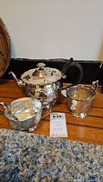 3 piece silver tone tea set displayed on floor with teapot, creamer, and sugar bowl showing detailed designs