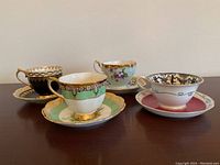 Four teacup and saucer sets displayed side by side on a wooden surface, showing varied patterns and colors including green, black, pink, and floral designs.