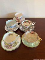 Five teacups and matching saucers arranged on a wooden surface showing various floral patterns and gold detailing.