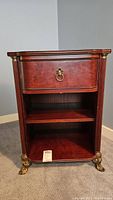 Front view of the Bombay night table with drawer closed and two open shelves visible. Shows reddish-brown wood finish with brass ring pull on drawer and clawed brass feet.