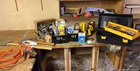 Workbench with various tools and small hardware supplies including a yellow toolbox with tools inside, drill bits in trays, containers of nails and staples, and painting supplies.
