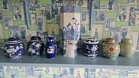 Seven assorted vases and ceramic jars displayed on a shelf against wallpaper. Various shapes and colors including dark blue and white floral jars, Asian vase, Wedgwood bud vase.