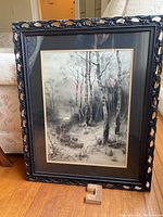 Full front view of framed black and white artwork showing sheep and trees in winter landscape with horse and herder, on wooden floor leaning against chair.